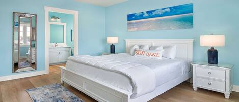A bedroom with a white bed, two bedside tables with lamps, a large mirror, a bathroom with a sink and cabinets, and a beach-themed painting on the wall.