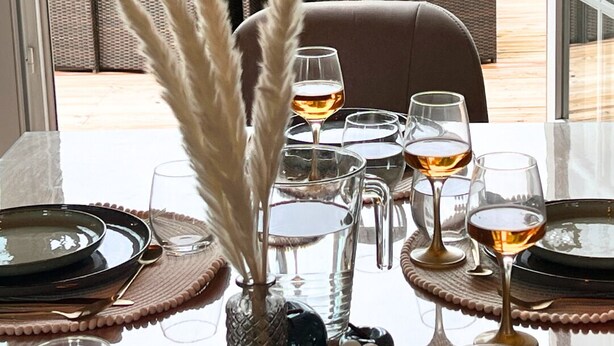 Une table à manger dressée avec des assiettes, des verres et un vase contenant des plumes d’herbe de la pampa.