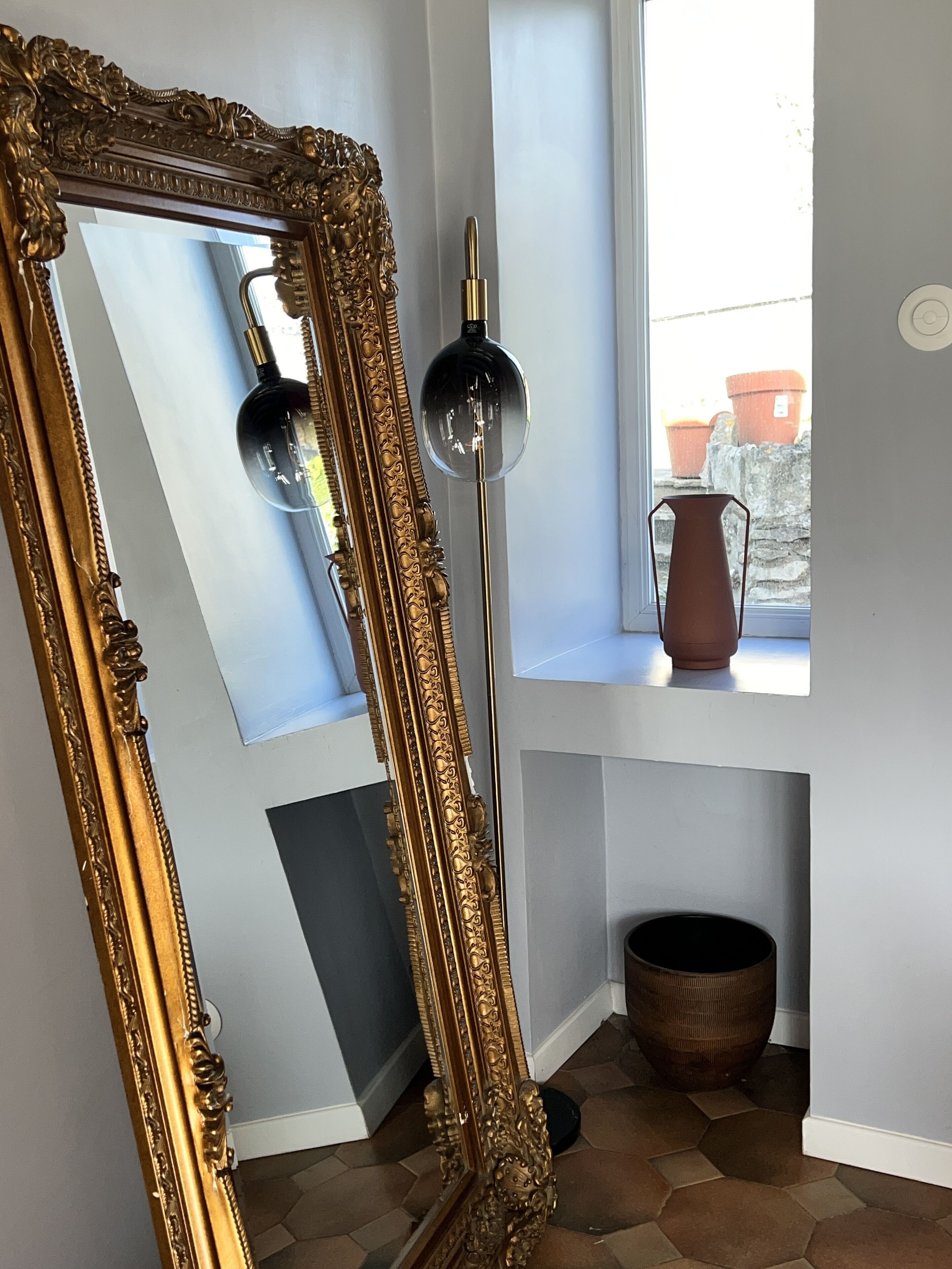 Une pièce comprenant un grand miroir décoratif appuyé contre un mur, un grand lampadaire, une fenêtre donnant sur l’extérieur et une petite table sur laquelle est posé un vase.