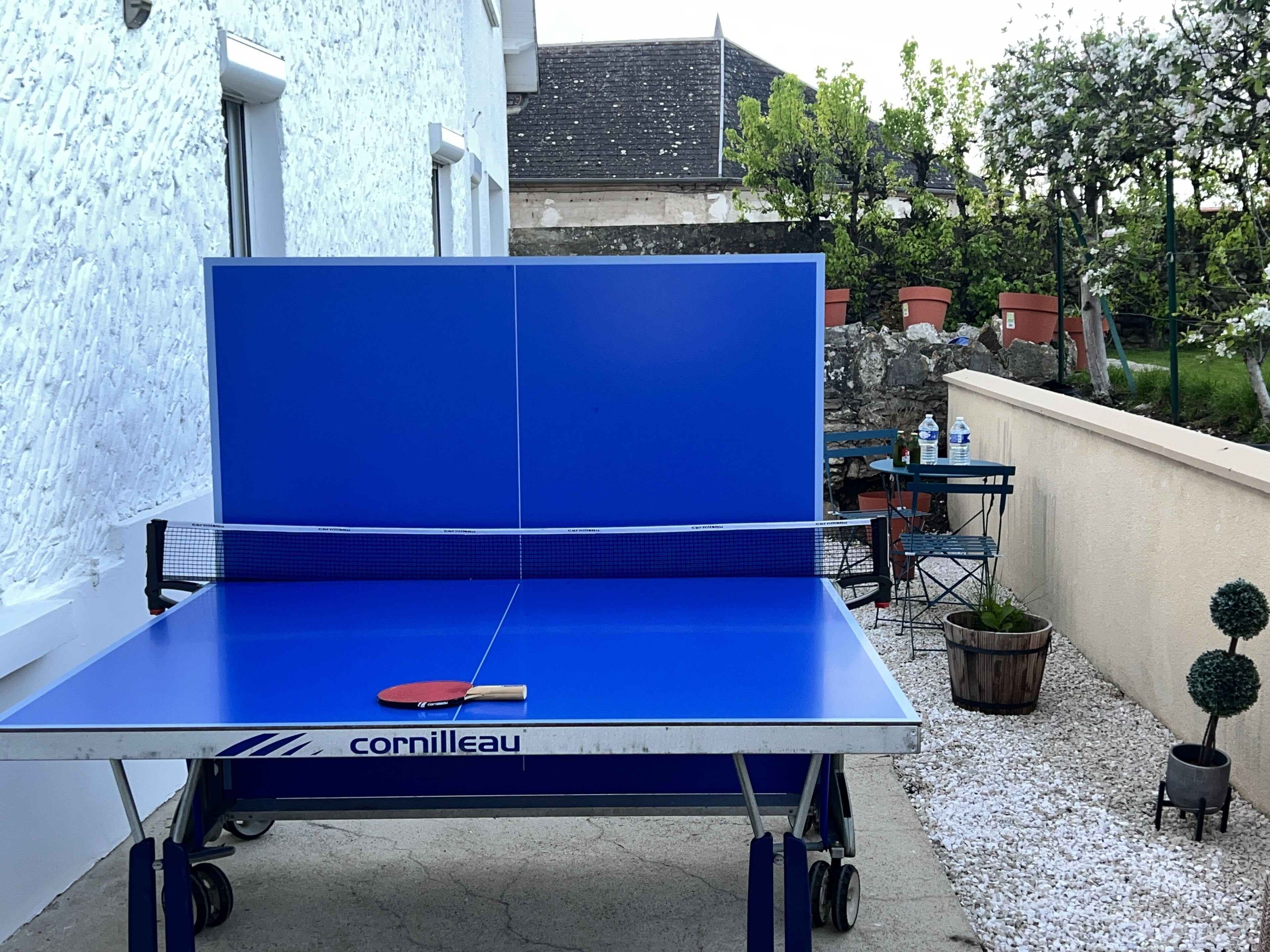 Een draagbare tafeltennistafel met een blauw speeloppervlak en een net, buiten geplaatst met een witte muur en een stenen muur op de achtergrond.