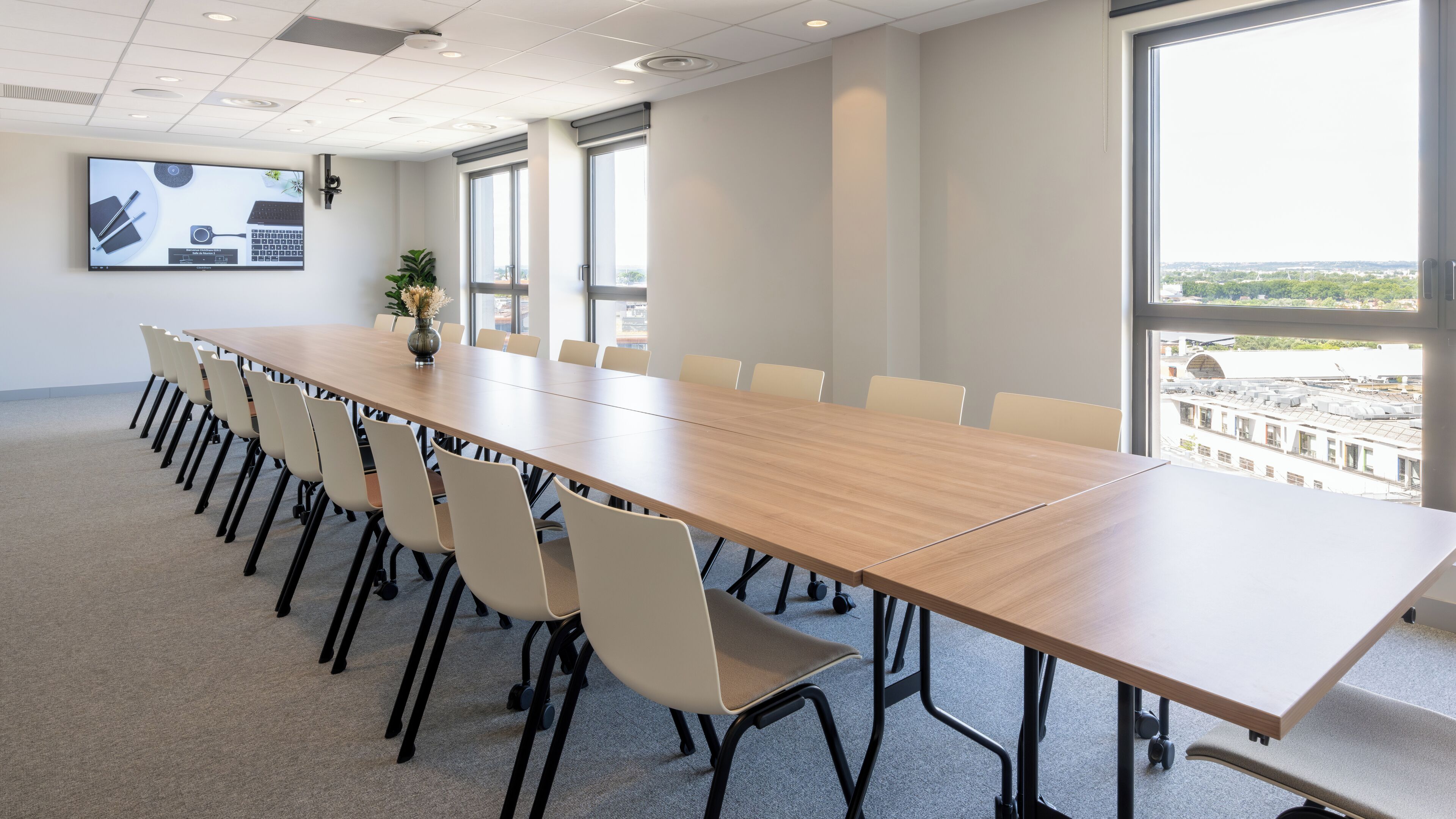 Un salón de conferencias con una larga mesa, sillas, una pantalla de proyector y ventanas amplias con vista a la ciudad.