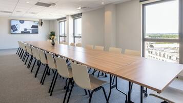 A conference room with a long table, chairs, a projector screen, and large windows with a city view.