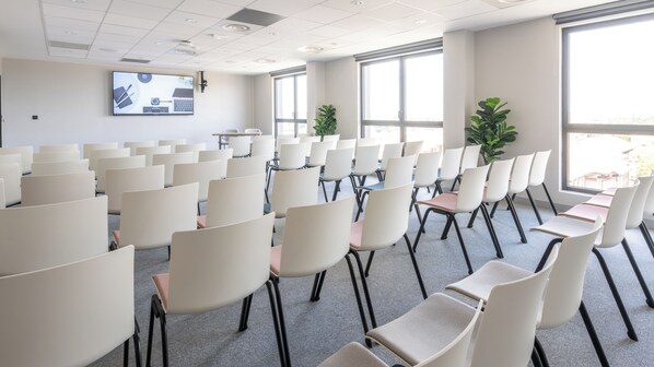 Une salle de conférence avec des rangées de chaises, un grand  écran et des fenêtres.