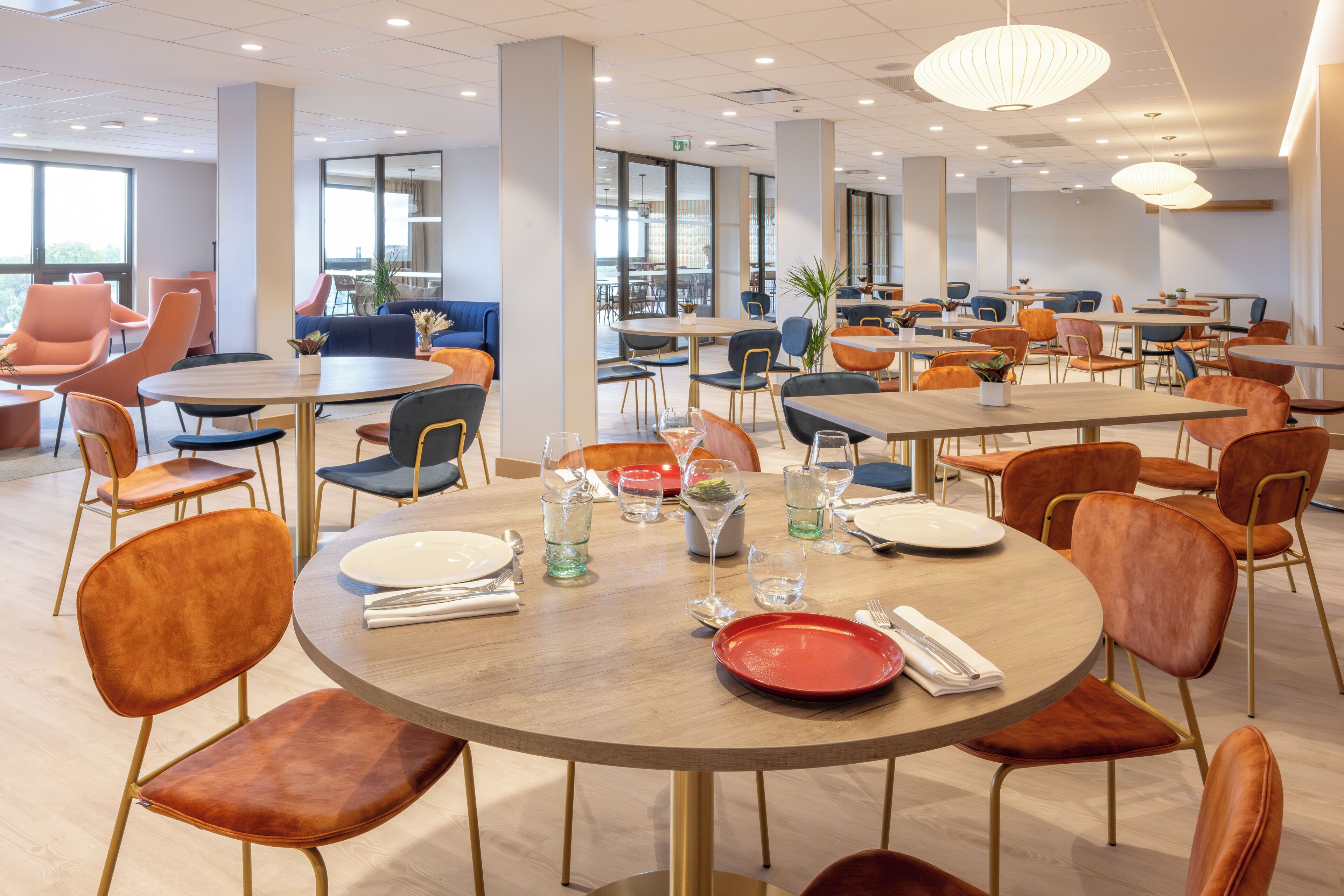 A spacious dining area with round tables, orange chairs, and modern pendant lighting.