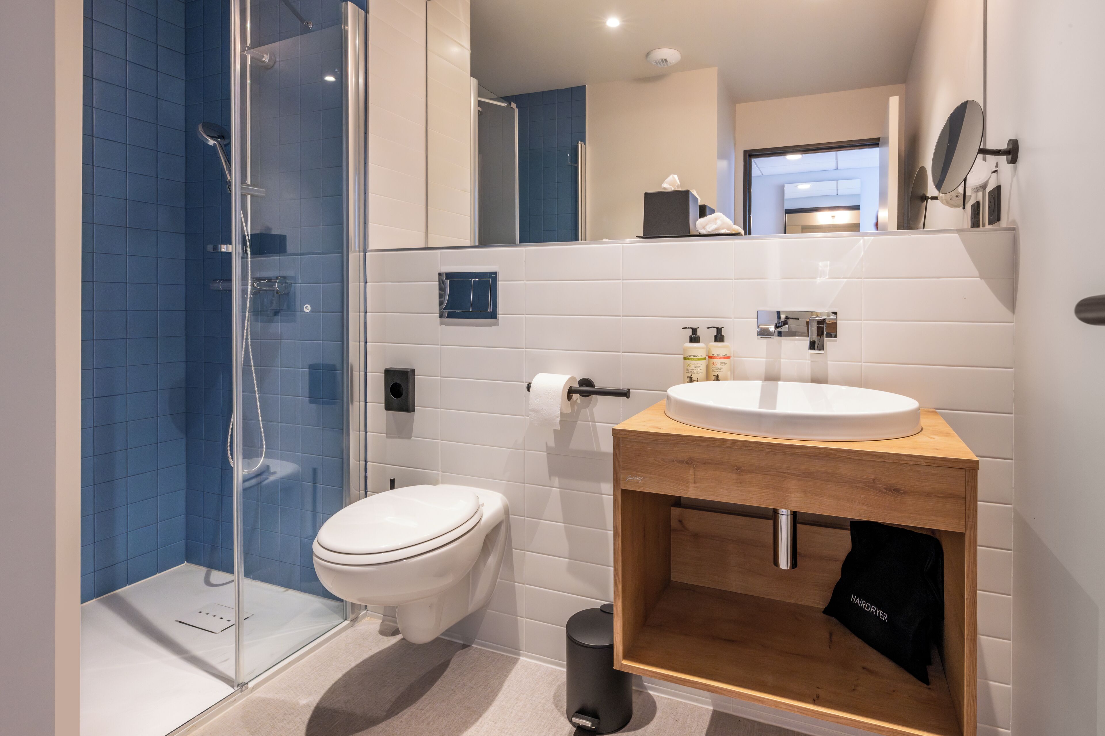 Et moderne badeværelse med en glas brusekabine, et hvidt toilet, et trækommode med vask og et spejl.