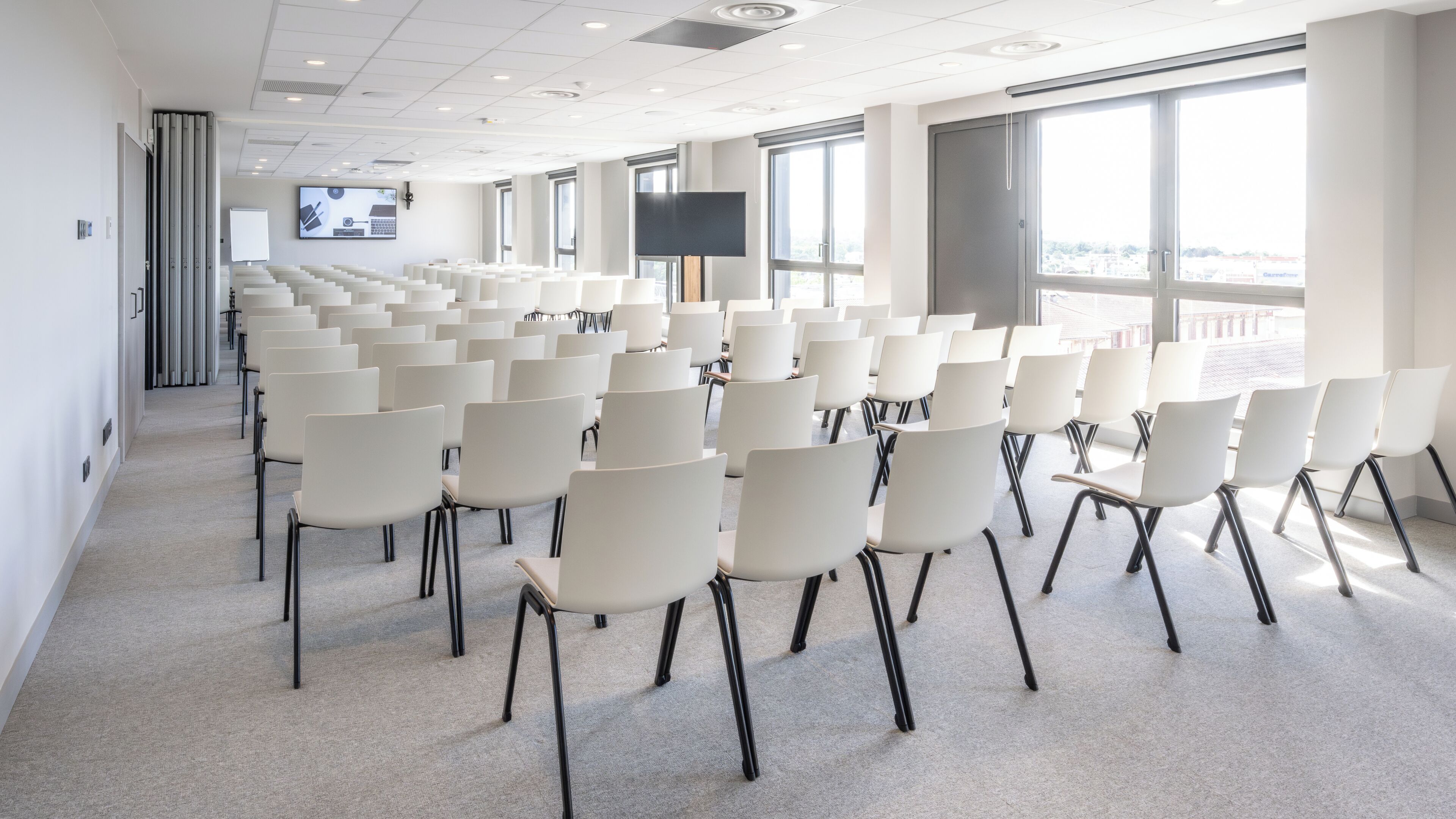 A conference room with rows of white chairs, large windows, and a flat-screen TV on the wall.