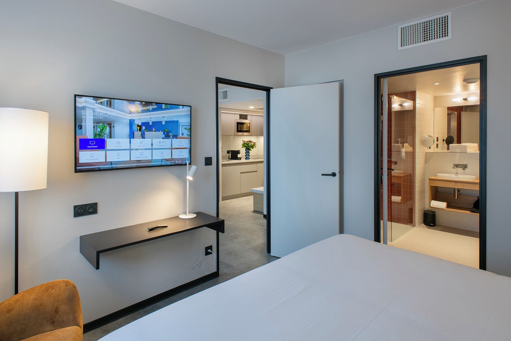 A modern hotel room with a bed, a TV mounted on the wall, a desk with a lamp, and a bathroom visible through an open door.