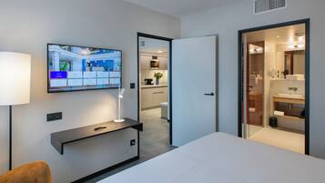 A modern hotel room with a bed, a TV mounted on the wall, a desk with a lamp, and a bathroom visible through an open door.