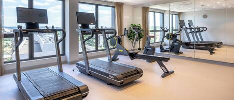 Een sportschool met loopbanden, hometrainers en een groot raam met uitzicht op de stad.