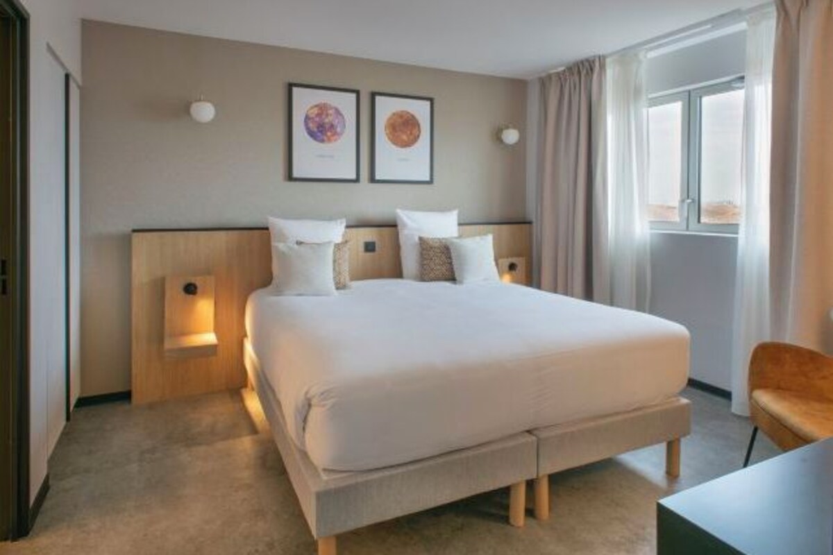 A hotel room with a large bed, two wall-mounted lamps, a desk, and a window with curtains.