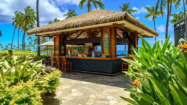 A thatched-roof bar with a stone floor, surrounded by tropical plants and palm trees, overlooking a beach. - Beach Villas at Ko Olina by KBM (Kapolei)