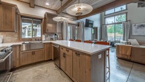 A spacious kitchen with wooden cabinets, a large island, and pendant lighting. There is a dining area with a table and chairs, and a living room with a sofa and a TV.