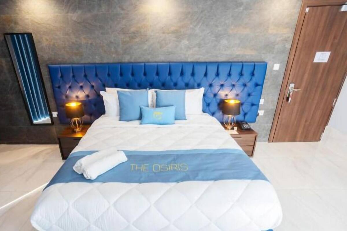 Une chambre d’hôtel avec un grand lit, une tête de lit capitonnée, deux tables de chevet avec des lampes et un mur d’accent bleu.