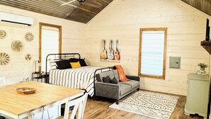 Een gezellige kamer met een houten plafond, een bed met streepjesbeddengoed, een grijze bank, een eettafel met stoelen en een klein bijzettafeltje met een plantje.