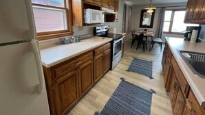 A compact kitchen with wooden cabinets, a refrigerator, a microwave, and a stove. There is a dining area with a table and chairs, and a window providing natural light. - R and M Escape (Detroit Lakes)