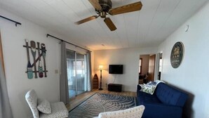 A living room with a ceiling fan, a blue sofa, two patterned armchairs, a TV, and a wall-mounted rack with paddles. - R and M Escape (Detroit Lakes)