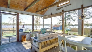 Una terraza de madera con un sofá, sillas y una mesa, con vista a un lago que tiene un muelle y un pequeño edificio al fondo.