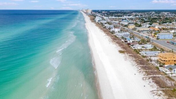 An aerial view of a coastal town with a long sandy beach, a dense cluster of buildings along the shore, and a clear blue sky.