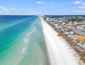 An aerial view of a coastal town with a long sandy beach, a dense cluster of buildings along the shore, and a clear blue sky. - Steps from the Beach! Your PCB Cottage Awaits! - E (Panama City Beach)