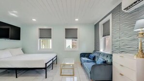 Een moderne slaapkamer met een bed, een slaapbank, een klein tafeltje en een dressoir. Er zijn twee ramen met zonwering, een aan de muur bevestigde televisie en een airconditioning.