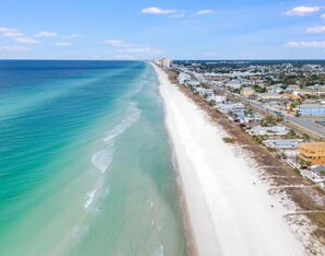 An aerial view of a coastal area with a long sandy beach, a road parallel to the beach, and a dense cluster of buildings. - Steps from the Beach! Your PCB Cottage Awaits! - C (Panama City Beach)