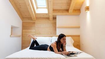A room with a sloped ceiling, wooden beams, a skylight, a bed with white bedding, and a wall-mounted light fixture.
