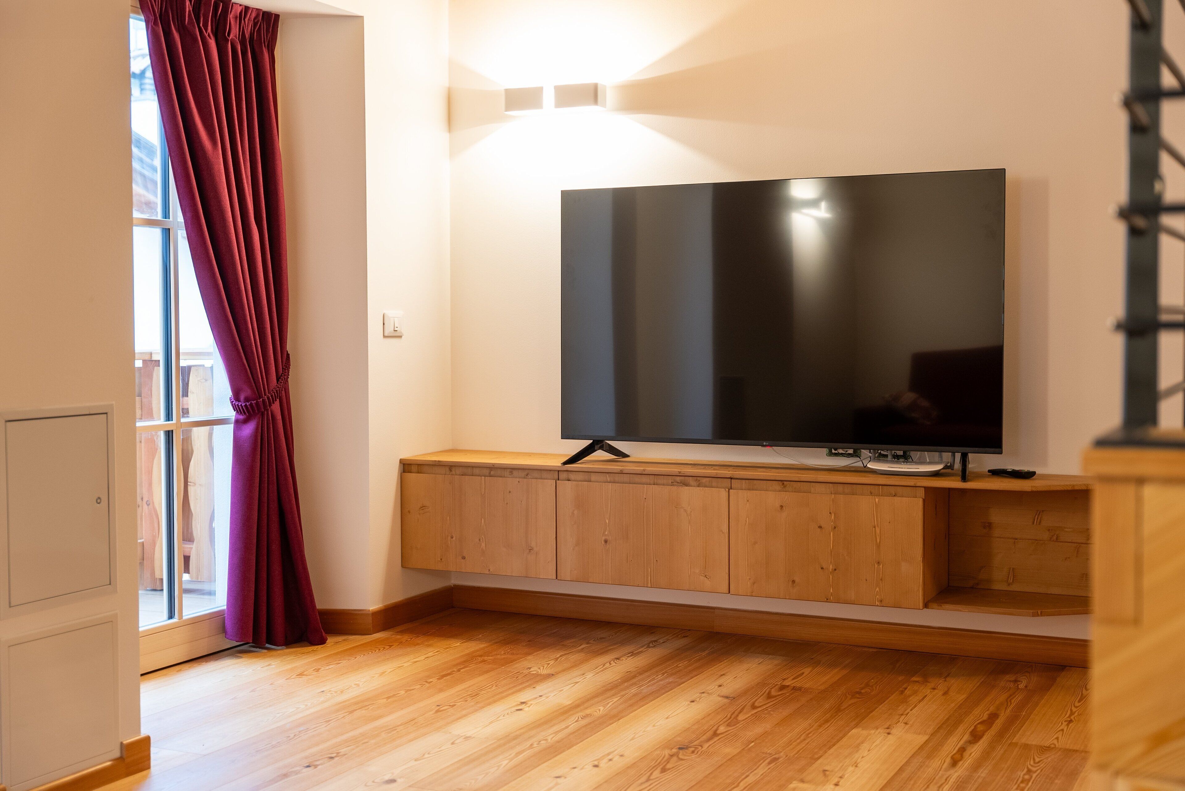 Een moderne woonkamer met een grote flatscreen-tv die aan een houten console is bevestigd, een rood gordijn en een houten vloer.