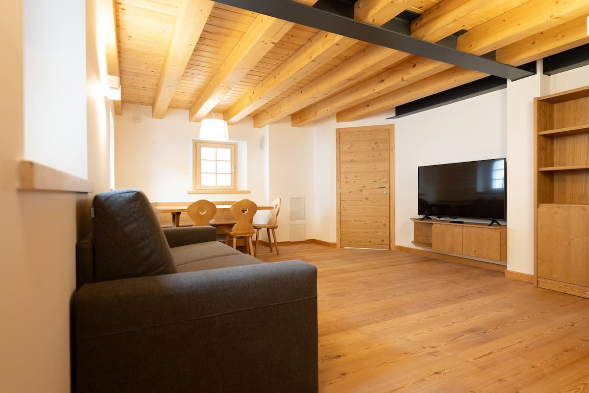 A modern living room with a sofa, wooden flooring, a TV mounted on a wooden cabinet, and a dining area with a table and chairs.