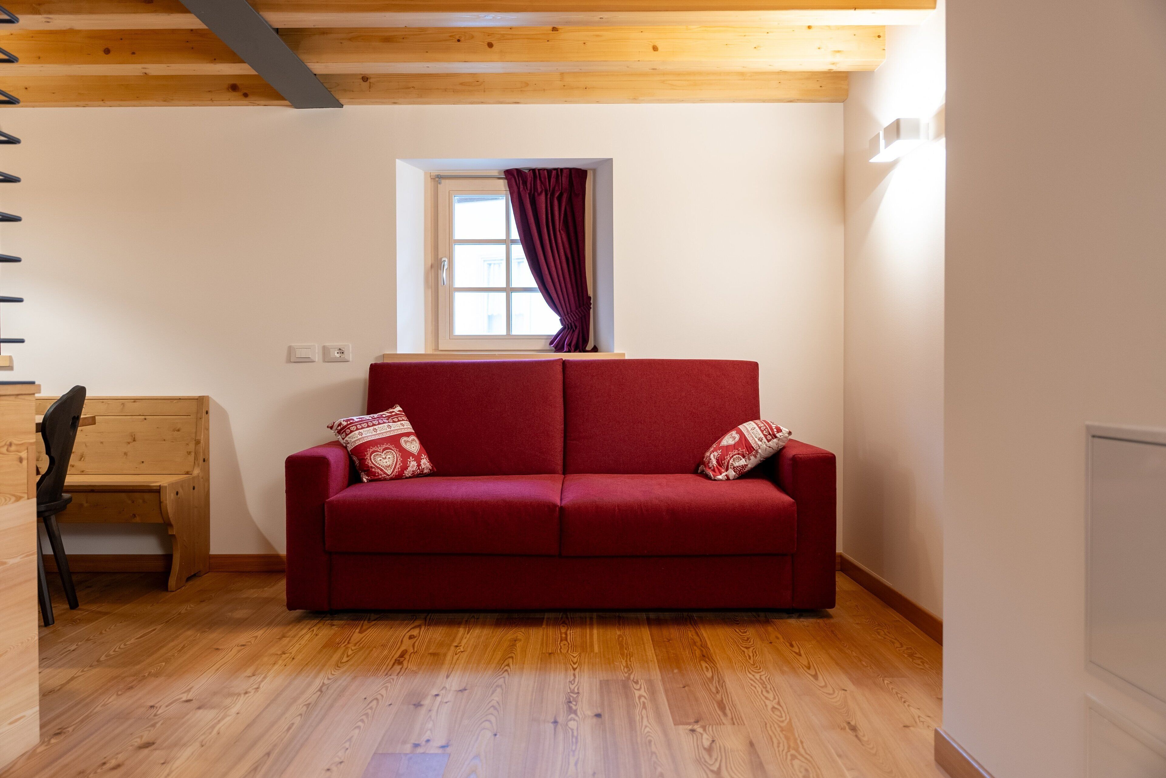 Un sofá rojo con cojines estampados en una habitación con suelo de madera y una ventana con cortinas.