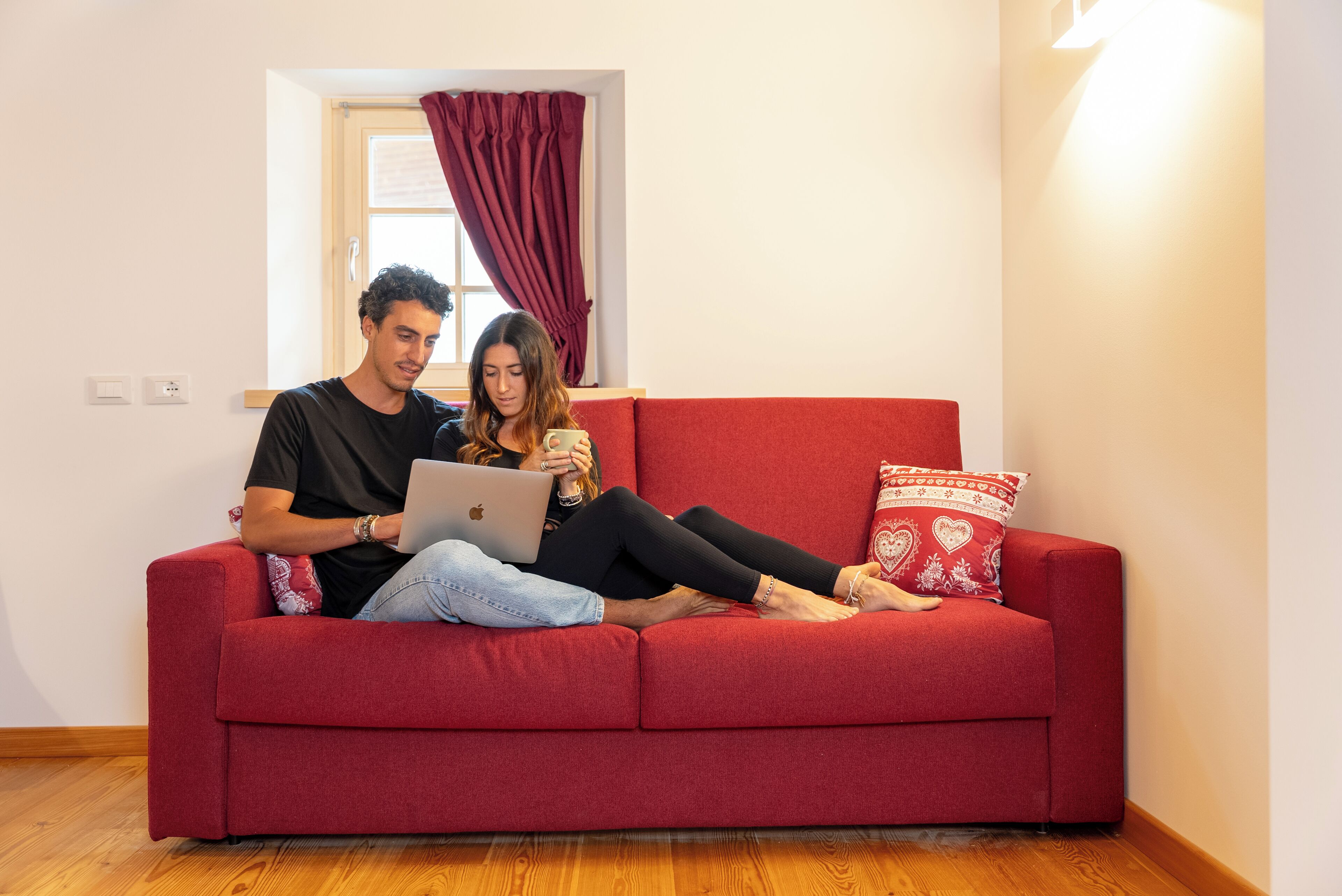 Un canapé rouge avec un ordinateur portable et une tasse, une fenêtre avec des rideaux et un luminaire fixé au mur.