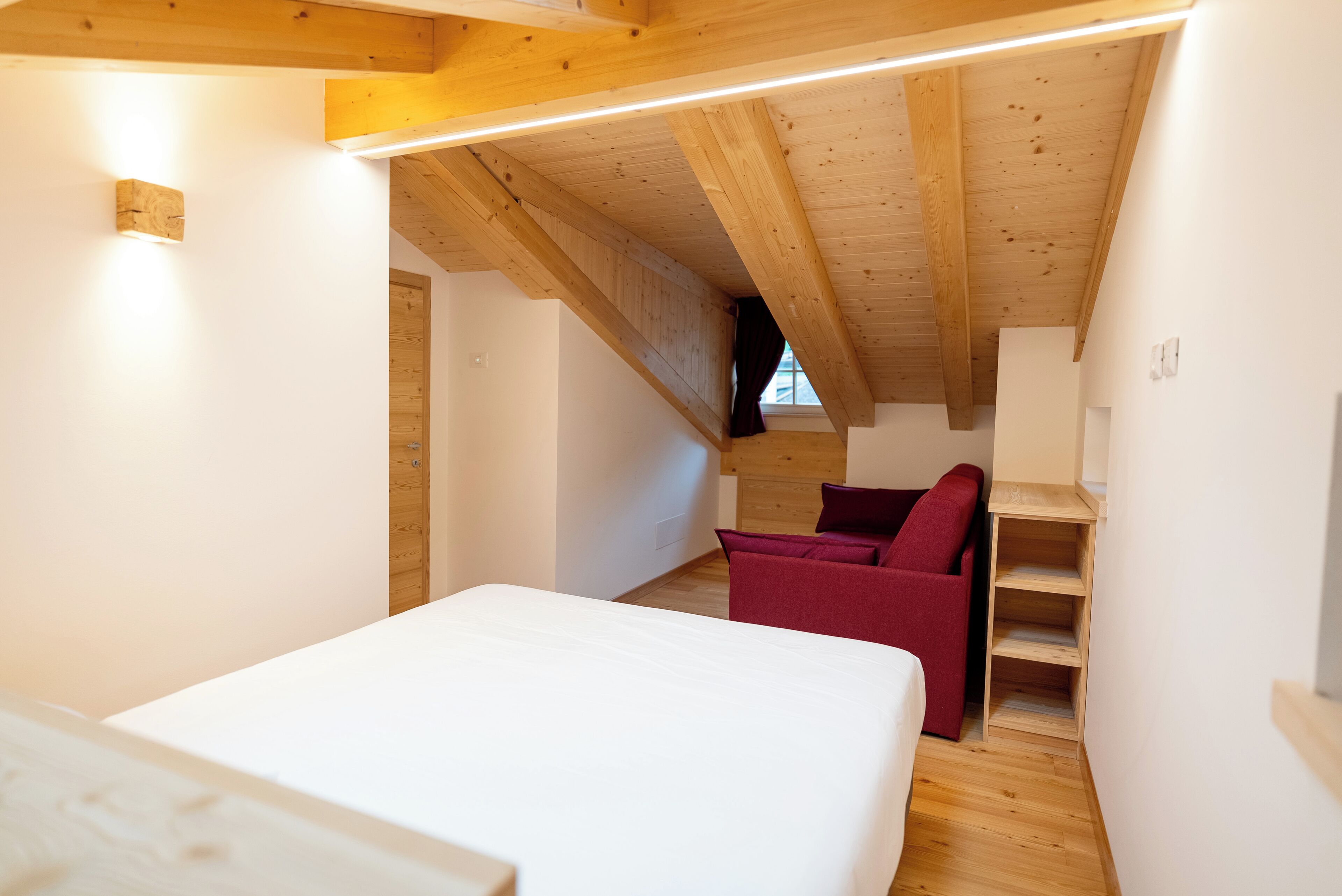 Une chambre avec un plafond en bois, un lit, une chaise rouge et une étagère en bois.