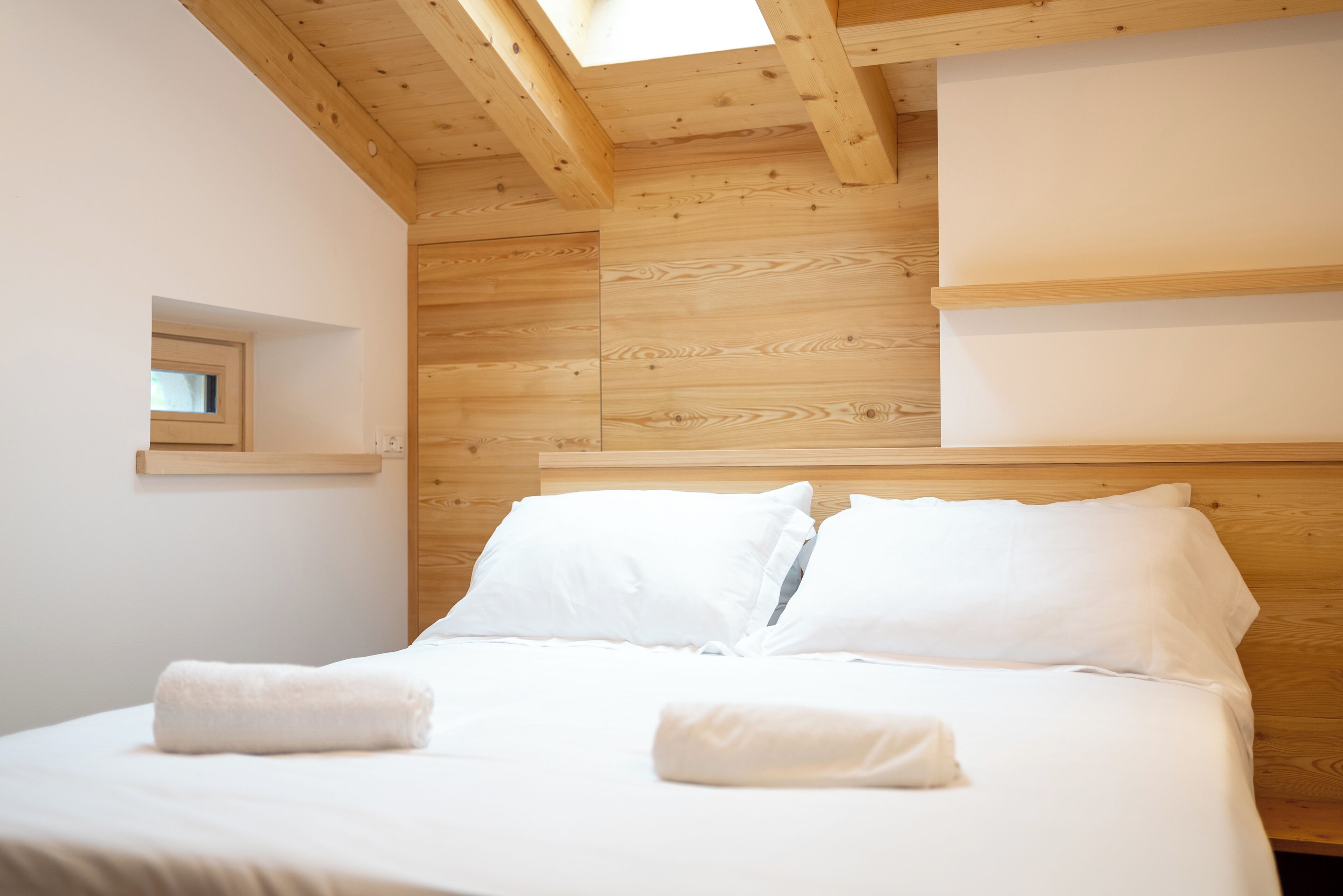 Un dormitorio con techo de madera, tragaluz, cama con ropa de cama blanca y una ventana pequeña.