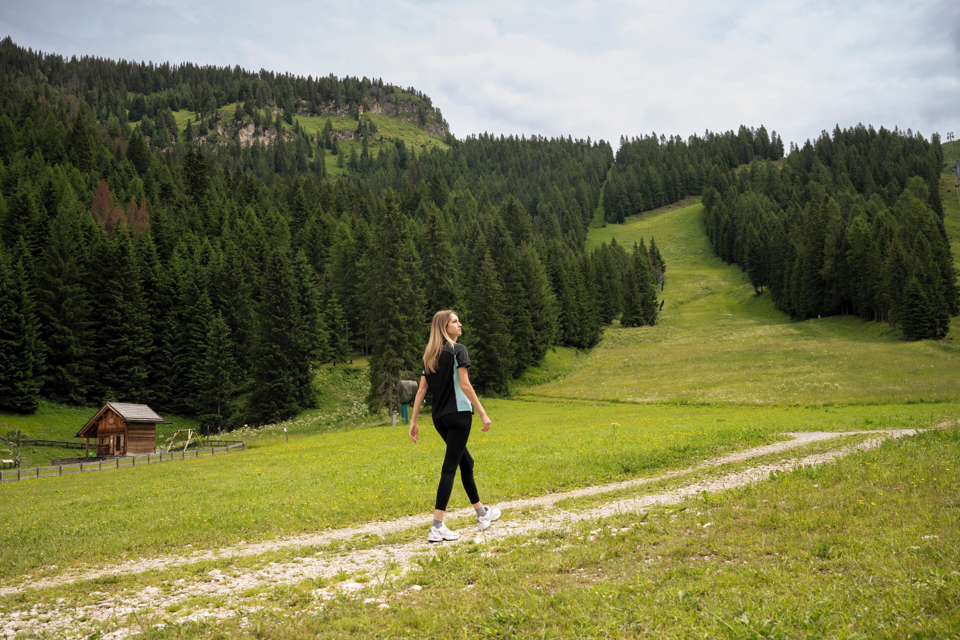 A person walking on a dirt path in a green, hilly area with a wooden cabin and dense forest in the background.