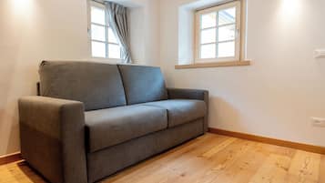 A grey sofa in a room with wooden flooring, two windows with curtains, and wall-mounted light fixtures.