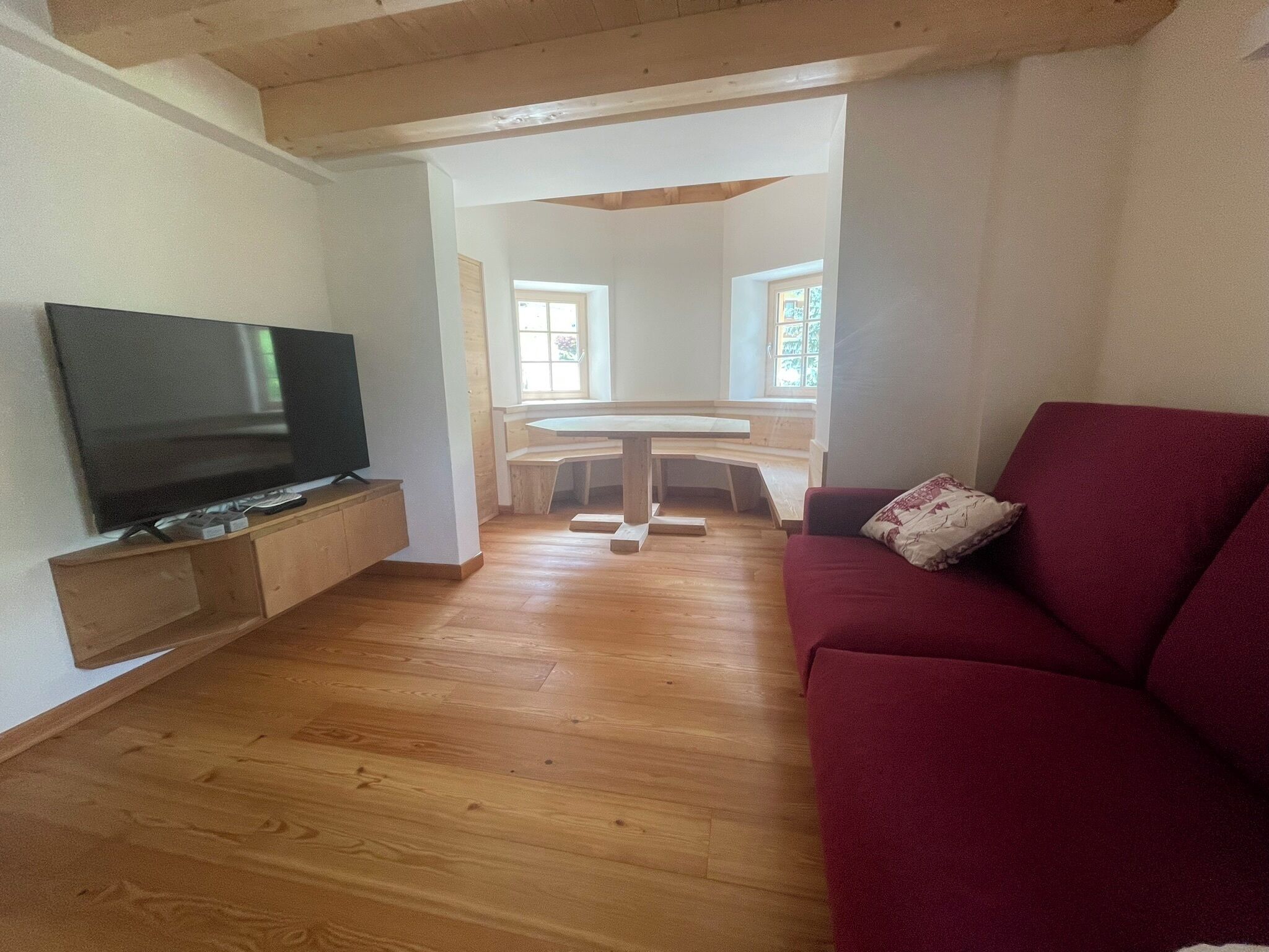 A living room with a TV, wooden flooring, a red sofa, and a dining area with a table and benches.