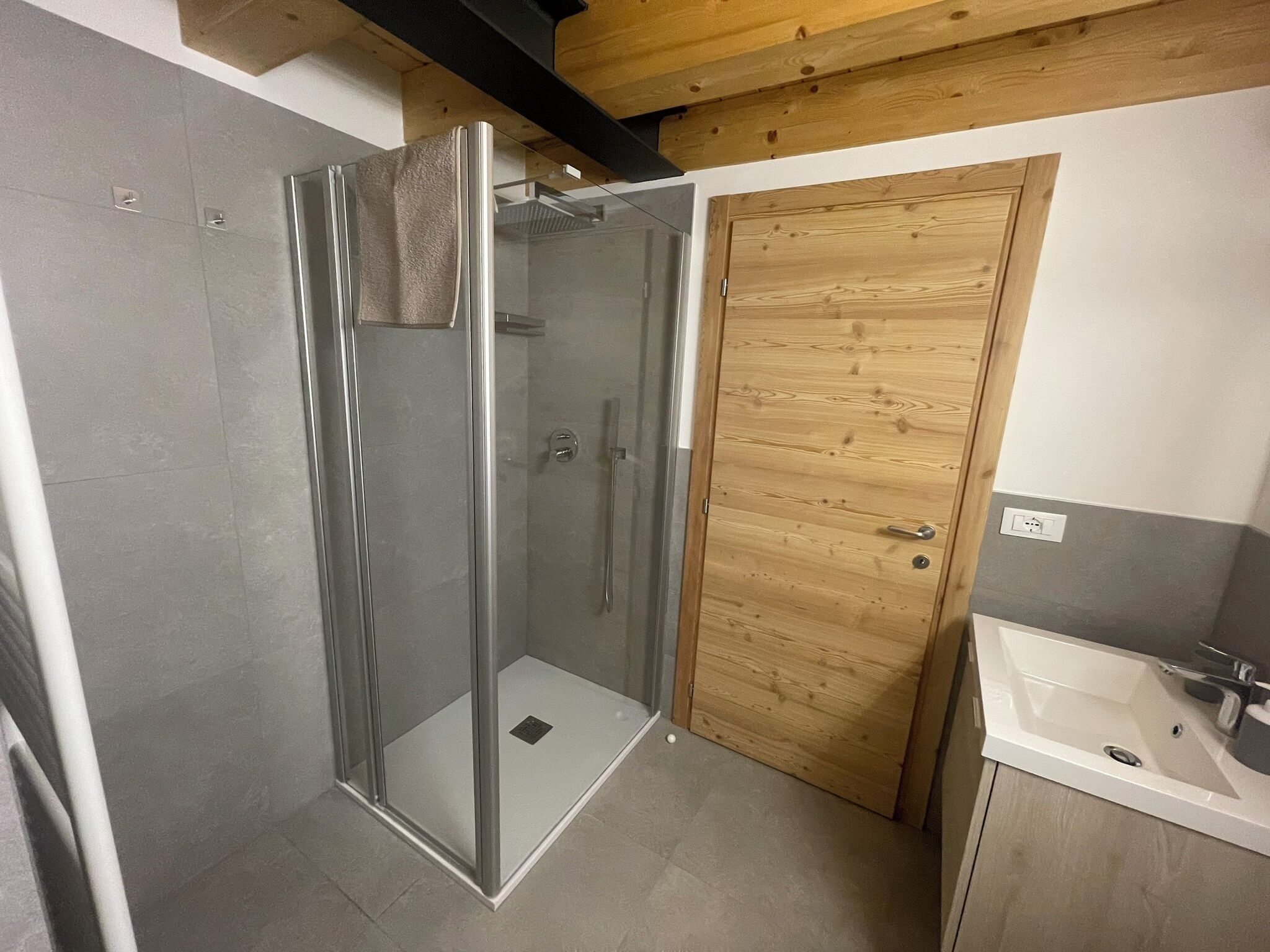 Un bany amb dutxa de vidre, una porta de fusta i un lavabo blanc.