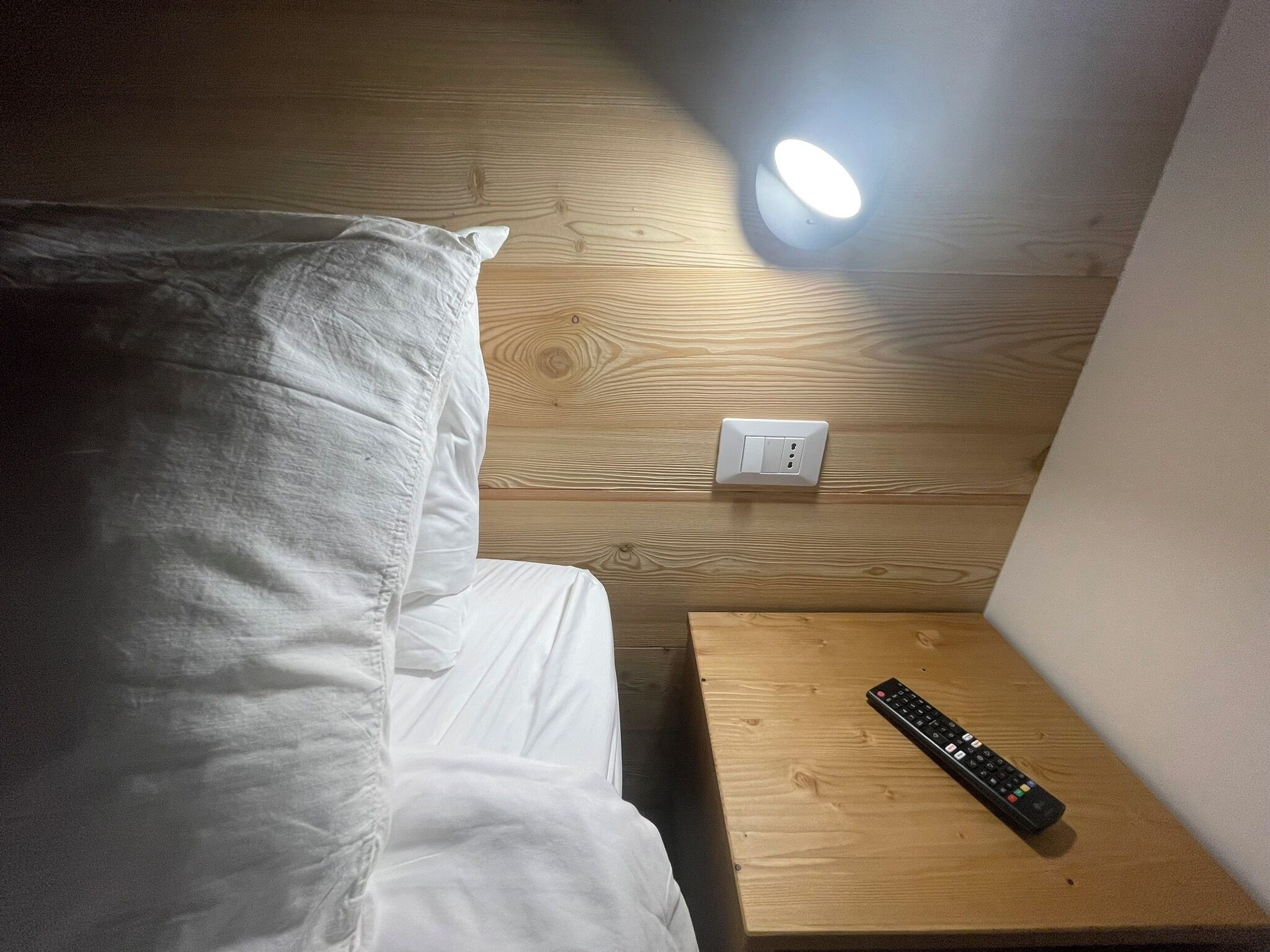 A hotel room with a wooden headboard, a white pillow, a wooden nightstand with a remote control, and a wall-mounted light fixture.