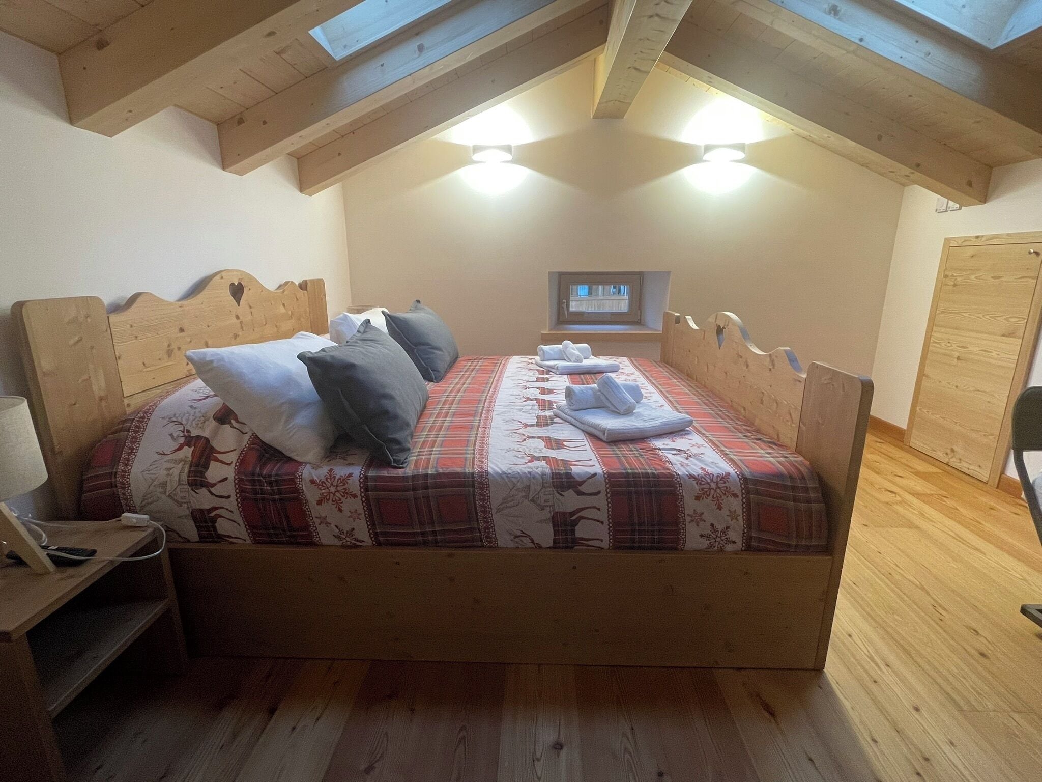Een houten bed met een geruit sprei en kussens, een nachtkastje met een lamp, en een raam met gordijnen.