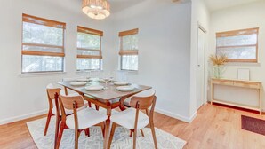 A dining area with a wooden table set for six, surrounded by six chairs. There are three windows with wooden blinds, a ceiling light fixture, and a small shelf with a vase and picture frame. - Hot Tub - E. Austin - 5 min to UT - 10 min to DT

 (Austin)