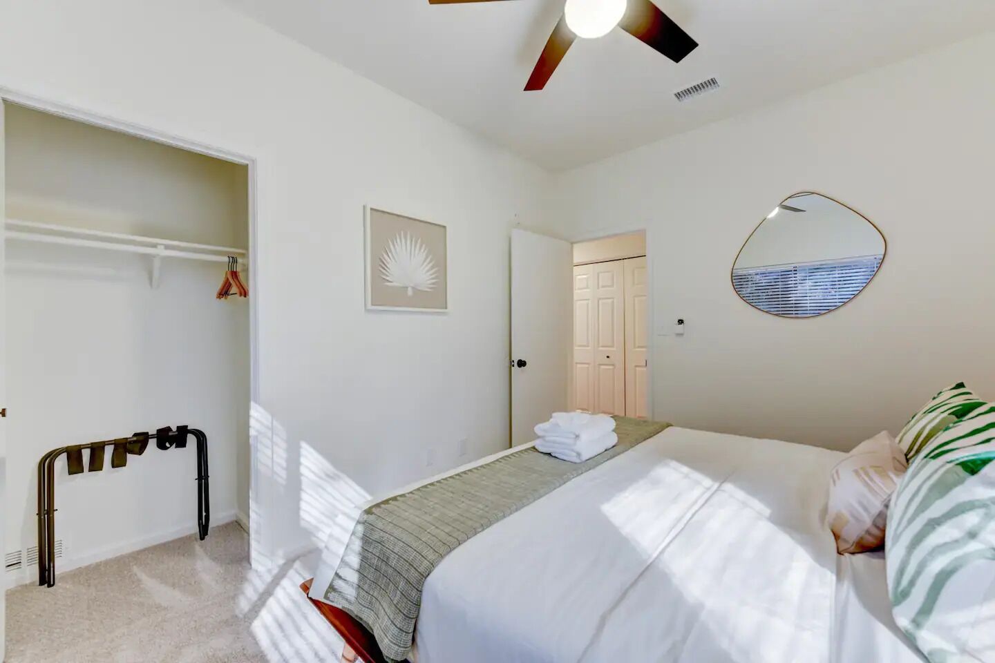 A bedroom with a bed, a bedside mirror, a ceiling fan, a door leading to another room, and a wall-mounted towel rack.