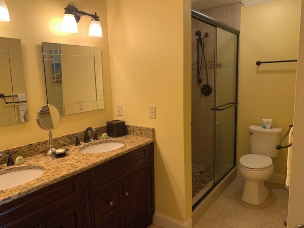 Baño con encimera de granito, lavabo doble, tocador, espejo y cabina de ducha con paredes de vidrio.