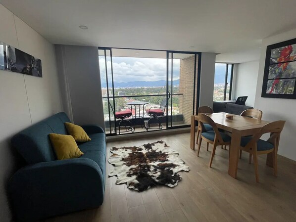 A modern living room with a blue sofa, a wooden dining table, and a large window offering a city view.