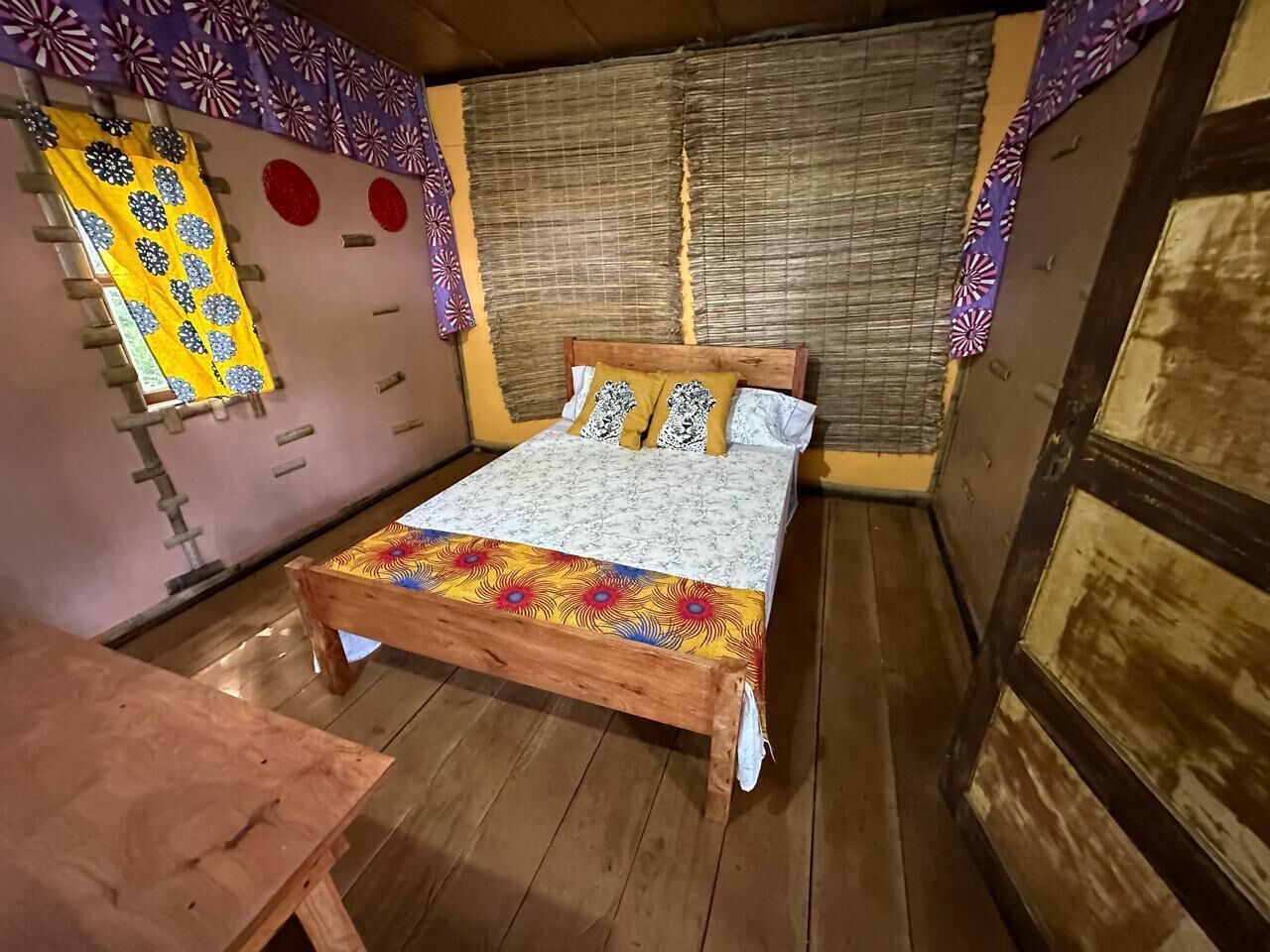 Uma cama de madeira com colcha estampada, um banco de madeira e uma janela com cortina.