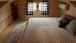 Een knus slaapkamer onder het dak met een houten schuine plafond, een bed met beddengoed met dessin, een klein houten kastje en een raam met geruit gordijnen.