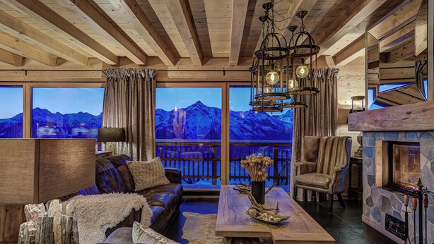 A cozy interior with a wooden ceiling, a large window overlooking mountains, a fireplace, and a seating area with a table and chairs. - Stunning Chalet Adele on the Piste (Veysonnaz)