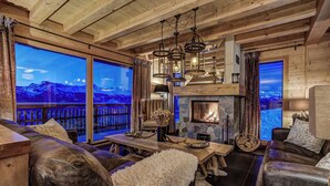 A cozy log cabin interior with a stone fireplace, leather furniture, and large windows overlooking a mountainous landscape. - Stunning Chalet Adele on the Piste (Veysonnaz)