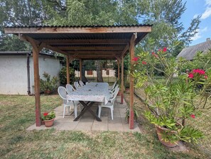 Een overdekte eethoek buiten, met een tafel en stoelen, omringd door potplanten en een tuin.