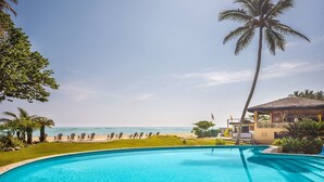 Piscina con vista a la playa y al océano, rodeada de palmeras y sillas de descanso.