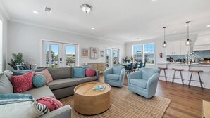 A spacious living room with a grey sofa, blue armchairs, and a round coffee table. A kitchen with white cabinets and a bar area is visible in the background. - Private Heated Pool, Fire Pit, 3 King Beds, 3 Min. Walk to the Beach, Dog Fri... (Gulf Shores)