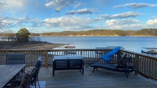 A wooden deck with a table and chairs, overlooking a lake with a dock and trees in the background. - Big fun lake at beaver lake (Rogers)